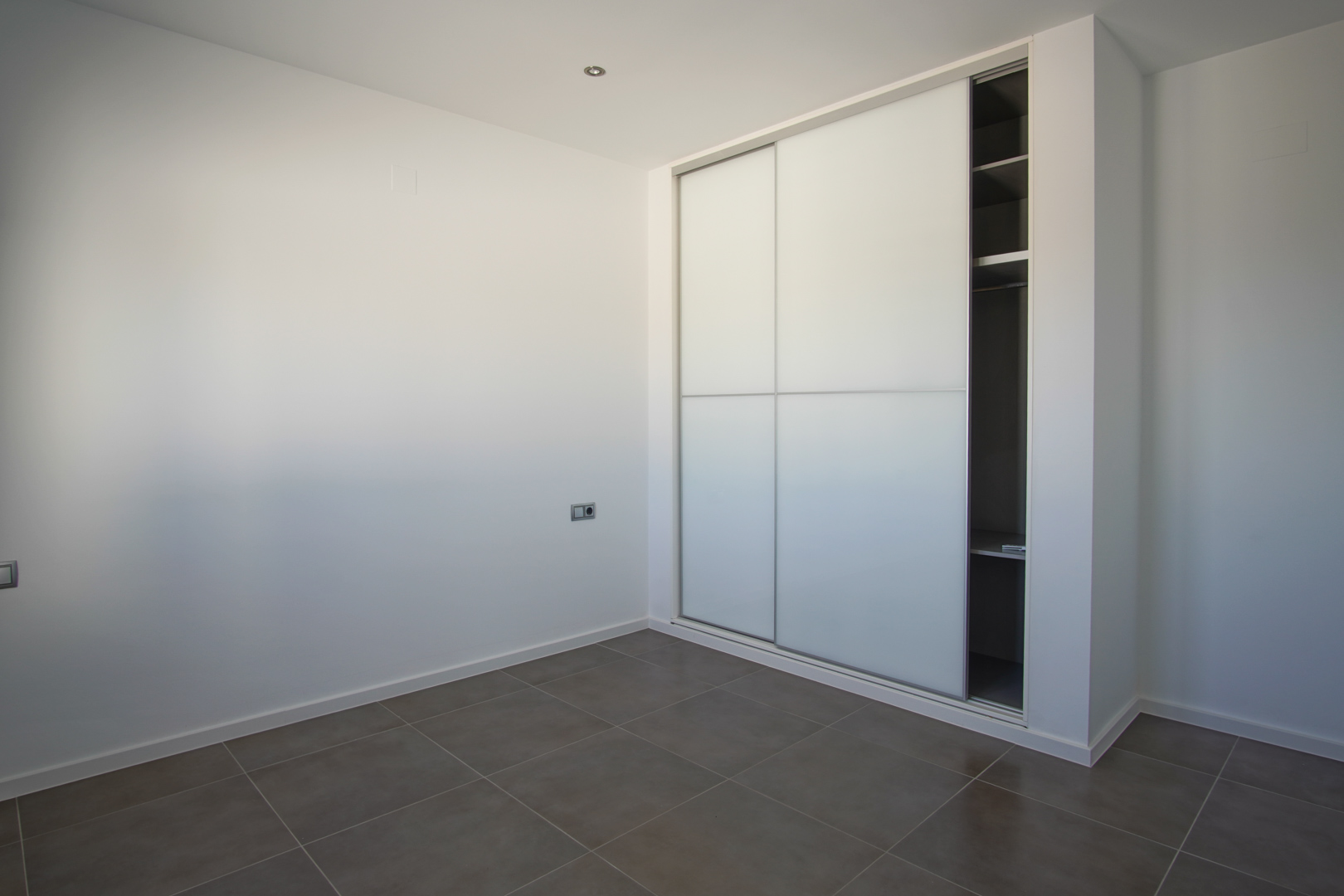 Einbauschrank mit weißen Schiebetüren und Inneneinteilung, kombiniert mit großformatigen grauen Bodenfliesen und minimalistischen weißen Wänden.
