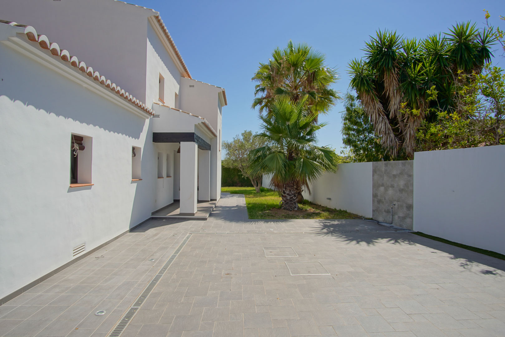 Gepflasterter Eingangsbereich mit grauen Fliesen, Bodenstrahlern und mediterraner Bepflanzung mit Palmen vor der weißen Fassade des Hauses.