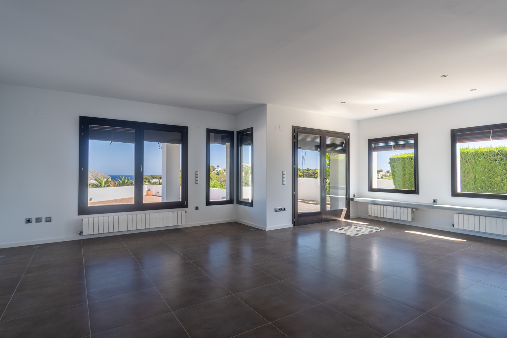 Großzügiger Wohnbereich mit dunklen Bodenfliesen, Heizkörpern und Holzoptik-Aluminiumfenstern mit weitem Blick auf das Meer und die Küste von Moraira.