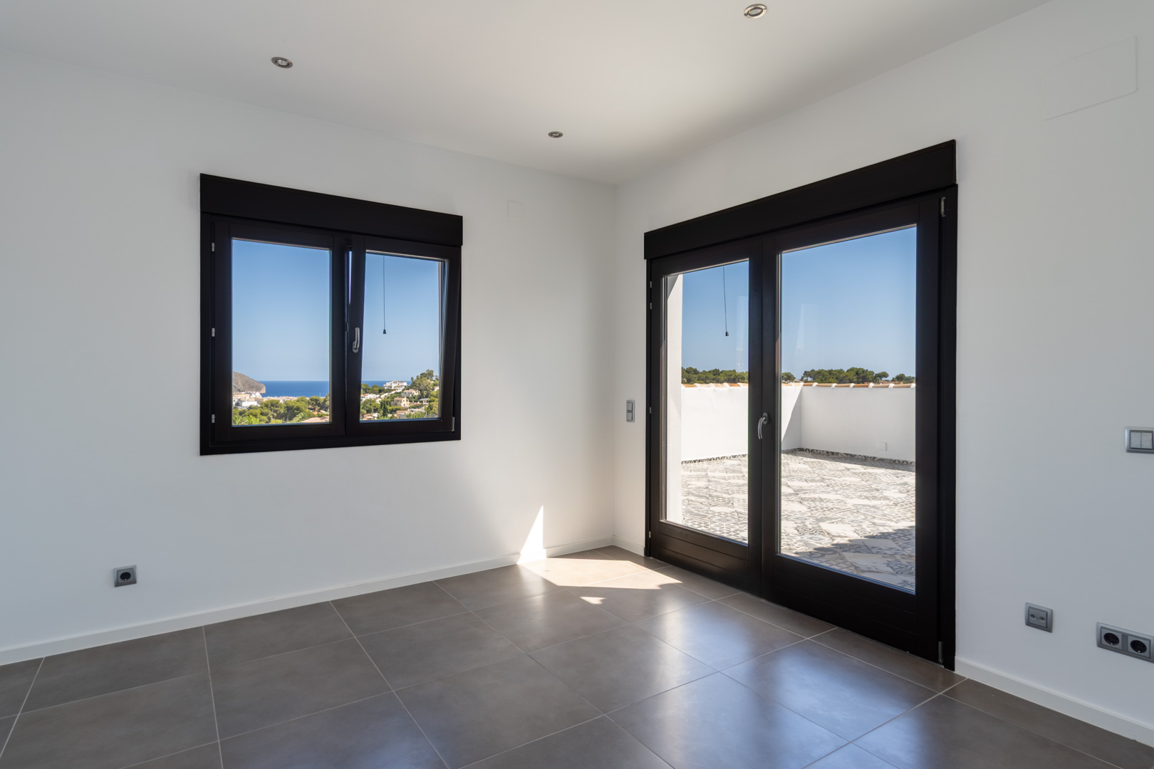 Heller Raum mit grauen Keramikfliesen, dunklen Fensterrahmen und direktem Zugang zur Terrasse mit weitem Blick auf das Meer und die Küste.