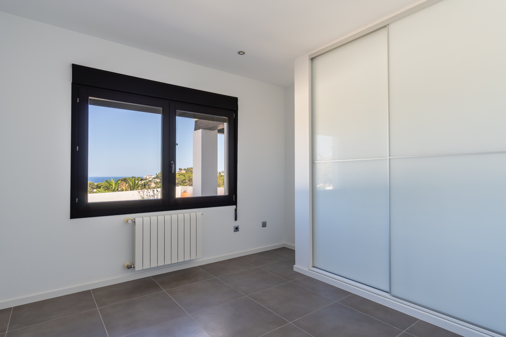 Helles Schlafzimmer mit Einbauschrank mit Schiebetüren, Heizkörper und dunklem Fensterrahmen mit Blick auf das Mittelmeer und die Küste von Moraira.