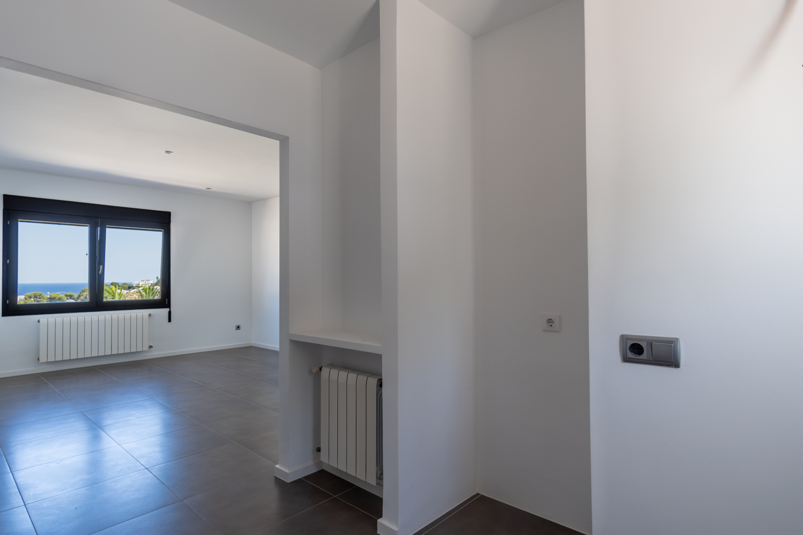 Innenansicht vom Flur mit grauen Keramikfliesen, Wandheizkörpern und einem schwarzen Aluminiumfenster mit Blick auf das Mittelmeer.