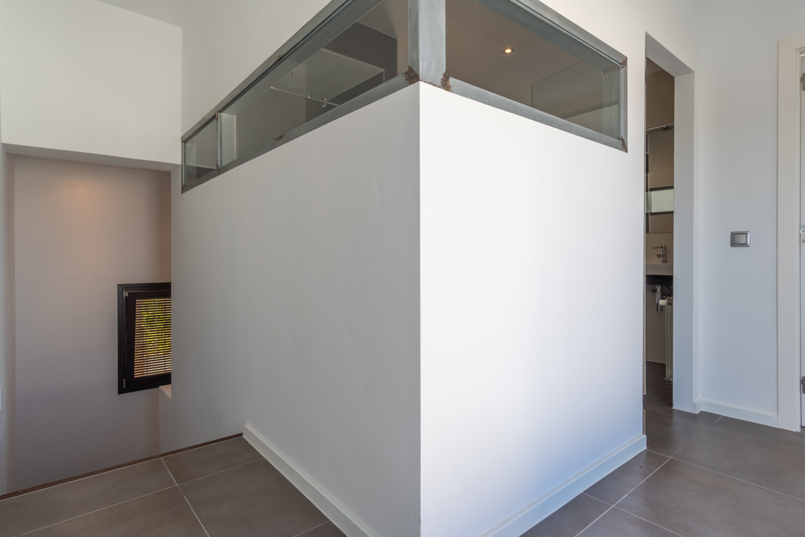 Innenflur mit grauen Bodenfliesen, Trennwand mit Glaselementen im Metallrahmen sowie Zugang zur Treppe und zum angrenzenden Badezimmer.