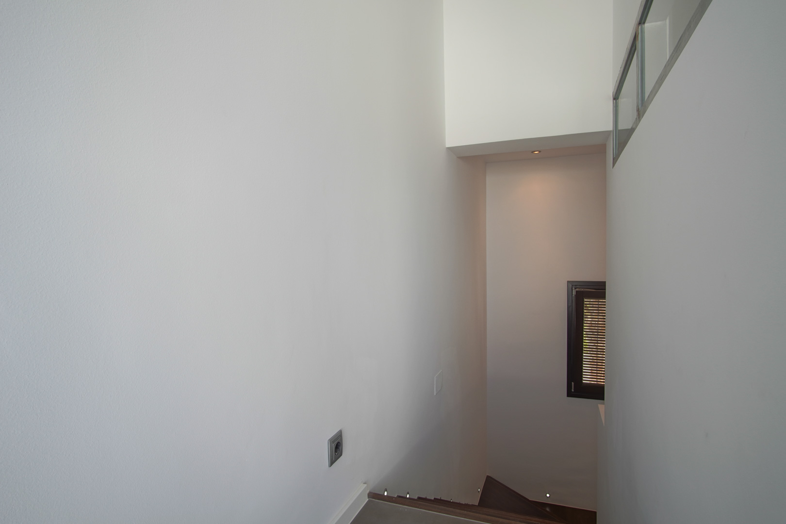 Innentreppe mit dunklen Stufen und integrierter LED-Bodenbeleuchtung. Schlichte weiße Wände und ein kleines Fenster mit dunklem Rahmen und Lamellenladen.