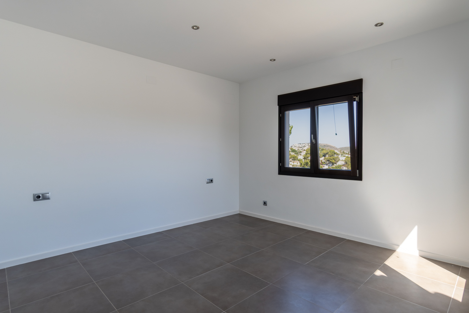 Minimalistisches Schlafzimmer mit grauen Feinsteinzeugfliesen, LED-Einbaustrahlern und dunklem Aluminiumfenster mit Blick auf die Umgebung.