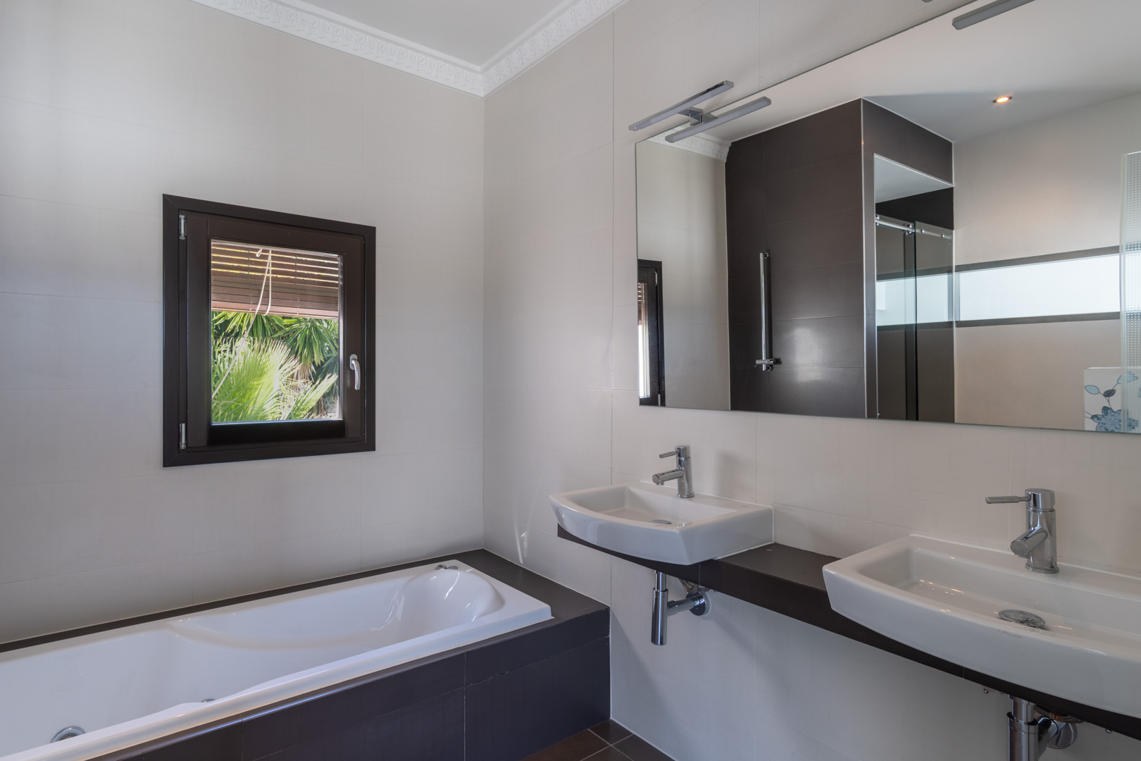 Modernes Badezimmer mit Doppelwaschbecken auf dunkler Konsole, Einbaubadewanne und Fenster mit Holzoptik-Rahmen für natürliches Licht und Gartenblick.