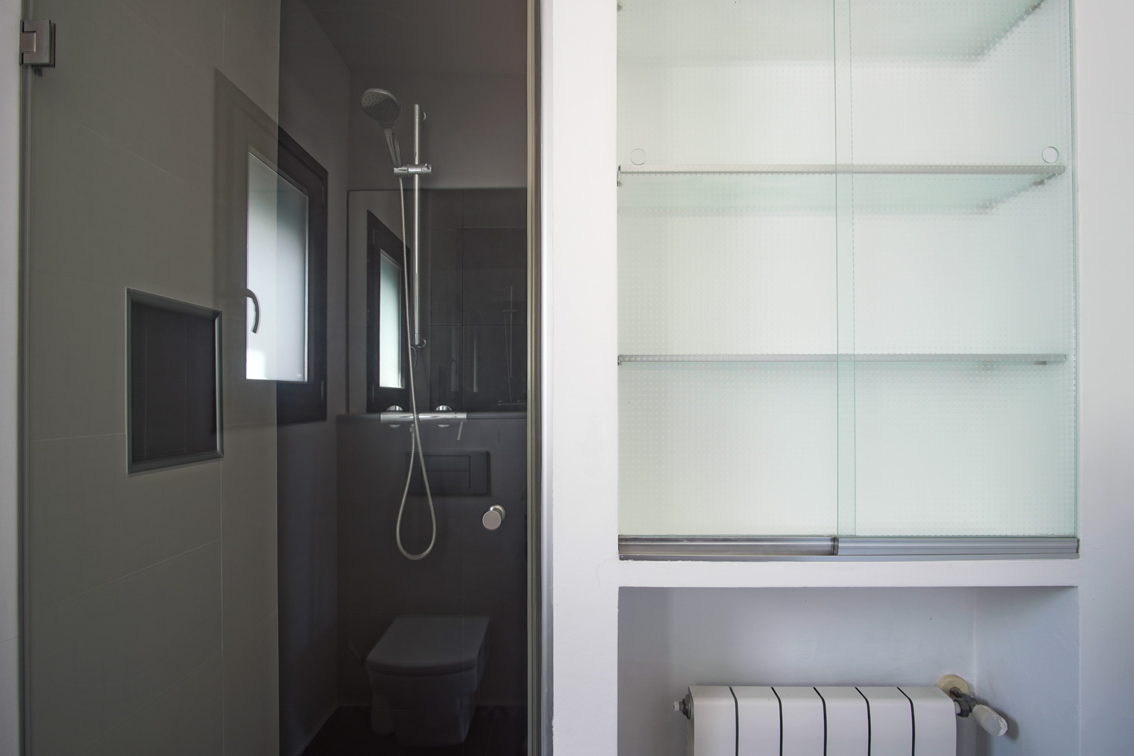 Modernes Badezimmer mit dunkelgrauen Fliesen, begehbarer Dusche und wandhängendem WC sowie einem Einbauschrank mit Glasböden und Heizkörper.