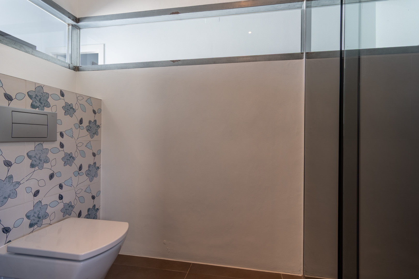 Modernes Badezimmer mit wandhängendem WC, dekorativen Fliesen mit blauem Blumenmuster und einem Oberlicht für natürlichen Lichteinfall.