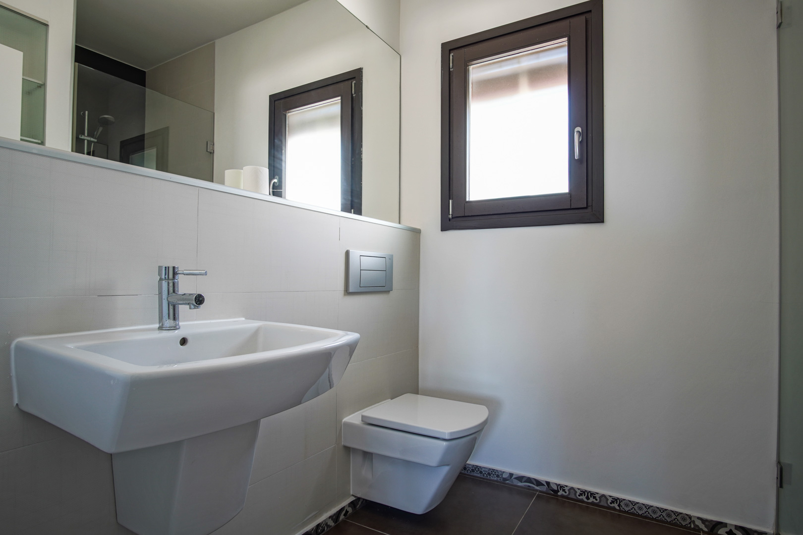 Modernes Badezimmer mit wandhängendem WC, weißem Keramikwaschbecken und Aluminiumfenster. Mit großem Spiegel und dunklen Bodenfliesen mit dekorativer Borte im hydraulischen Stil.