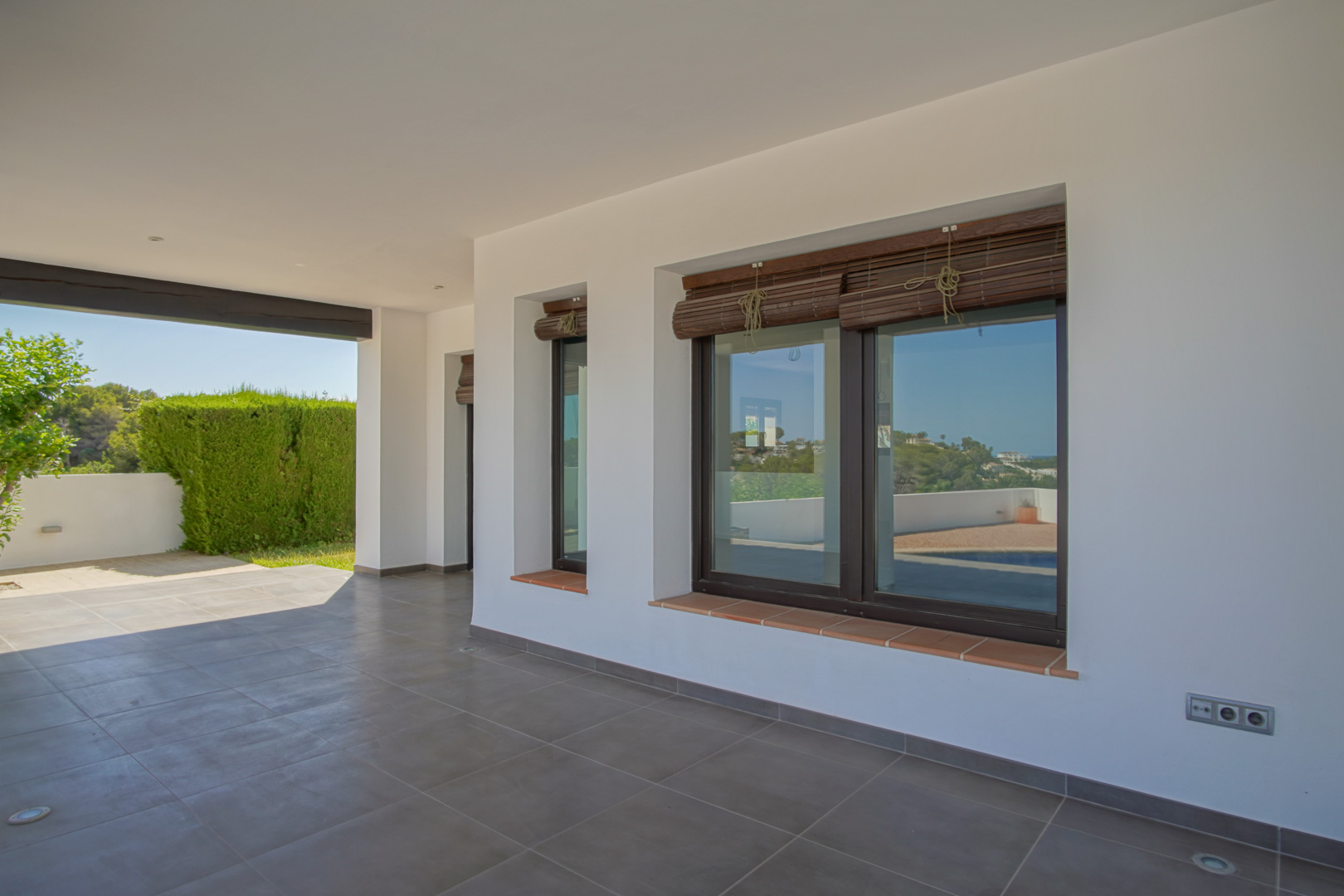 Überdachte Veranda mit grauen Bodenfliesen, weißen Wänden und Aluminiumfenstern in Holzoptik mit Bambusrollos und Fensterbänken aus Terrakotta.