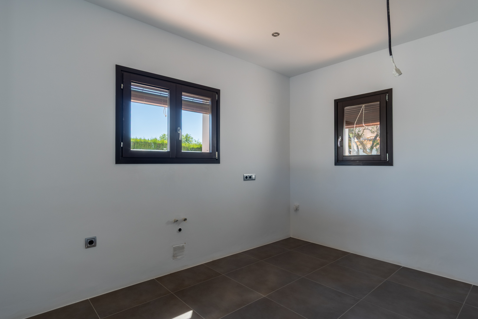 Vorbereiteter Raum für die Kücheninstallation mit dunklen Bodenfliesen, Aluminiumfenstern in Holzoptik und vorhandenen Sanitär- sowie Elektroanschlüssen.