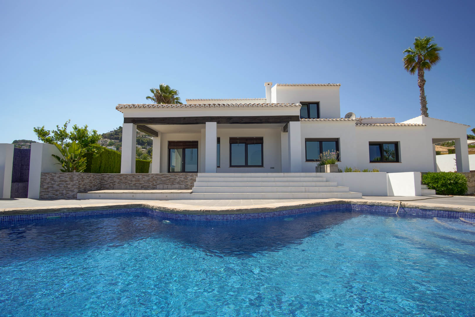 Weiße mediterrane Villa mit blauem Mosaikpool. Die Außenansicht zeigt eine überdachte Terrasse mit dunklen Holzbalken und breiten Steinstufen zum Wohnbereich.