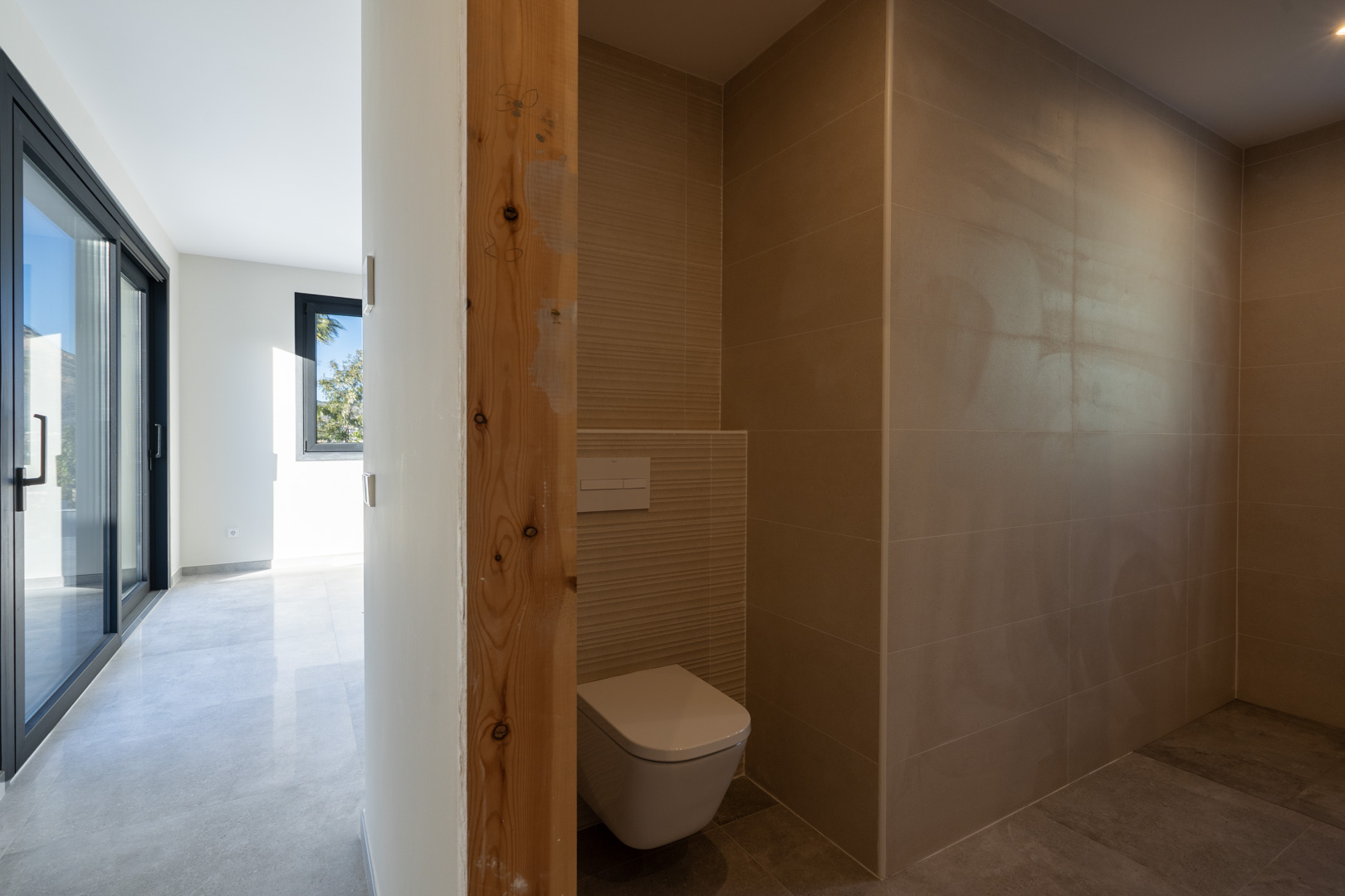 Detail eines Gäste-WC mit sandfarbenen Fliesen und rohem Holztürrahmen mit Blick auf den Flur.