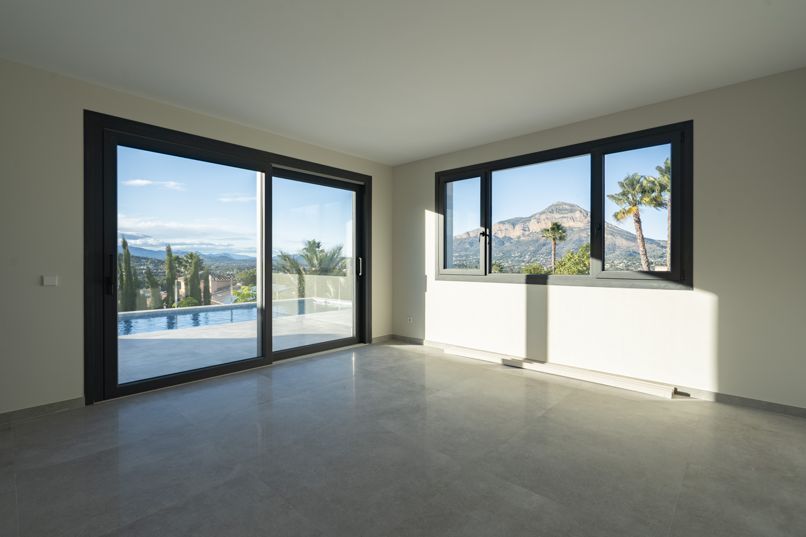 Geräumiges Wohnzimmer mit grauem Fliesenboden und großen Fenstern mit Blick auf den Pool und die Berglandschaft.