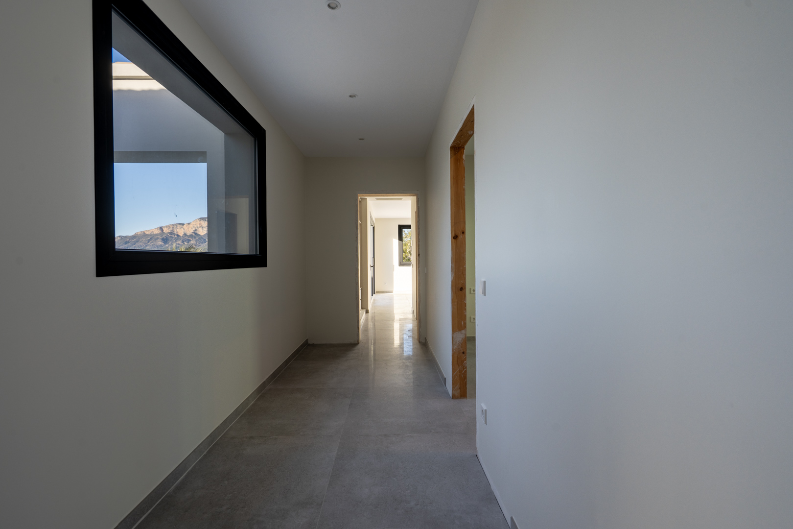 Heller Flur mit grauem Fliesenboden und Bergblick durch ein rechteckiges Fenster mit schwarzem Rahmen.