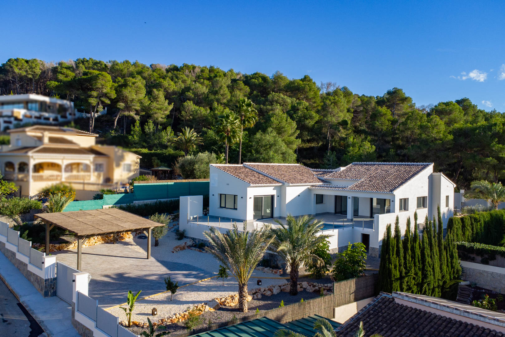 Luftaufnahme einer modernen Villa mit braunen Dachziegeln, weißer Fassade und überdachtem Carport in Jávea, umgeben von Grün.