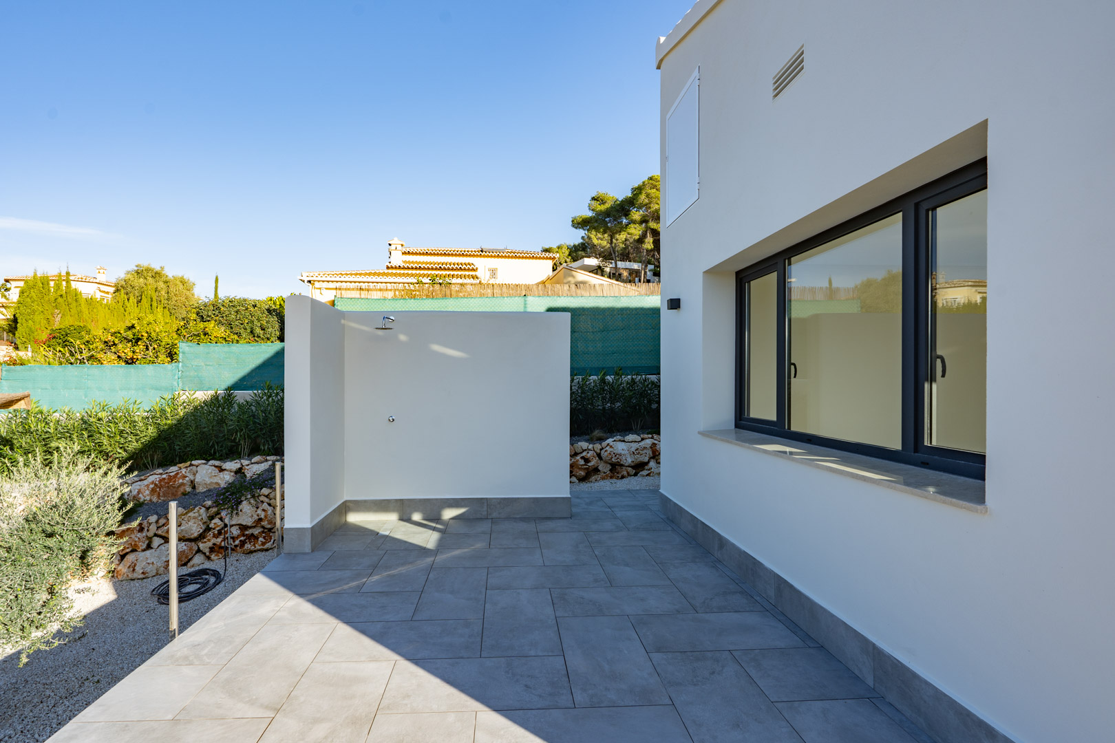 Patio mit Außendusche und grauen Fliesen, neben einer weißen Wand mit doppelt verglastem Fenster.