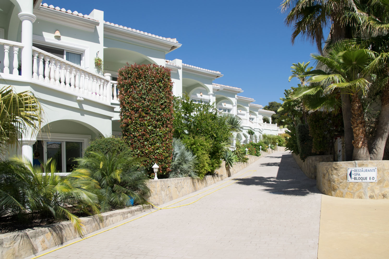 Außenansicht der mediterranen Anlage mit weißen Balustraden, Bogenterrassen und gepflegten Gärten entlang eines hellen, gepflasterten Zugangsweges.