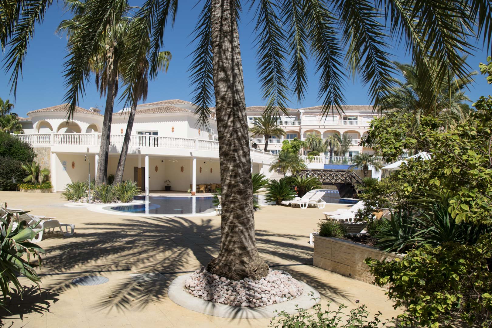 Außenbereich mit Lagunenpool, umgeben von Palmen und mediterraner Bepflanzung. Die Architektur umfasst weiße Fassaden, Rundbogen-Terrassen und weitläufige Liegeflächen.