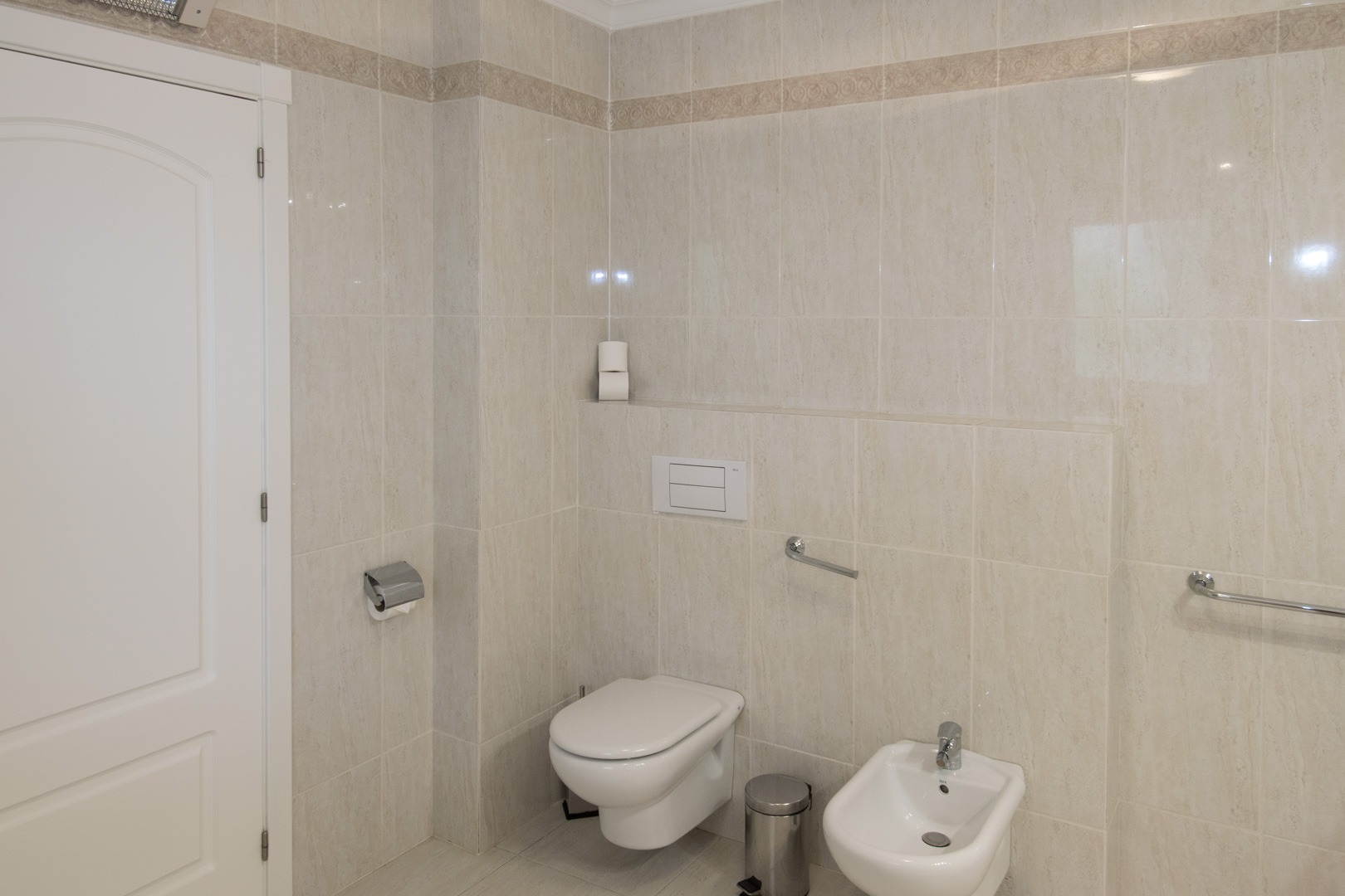 Badezimmer mit cremefarbenen Fliesen und Zierbordüre, ausgestattet mit Hänge-WC, Bidet und verchromten Sicherheits-Haltegriffen.