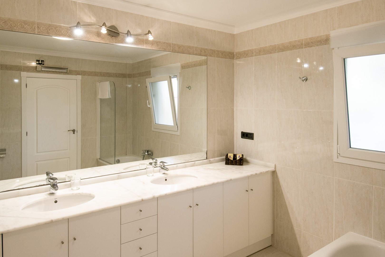Badezimmer mit Doppelwaschbecken auf einer weißen Marmorplatte und großem Wandspiegel. Die Wände sind mit beigefarbenen Fliesen und einer dekorativen Bordüre gestaltet.