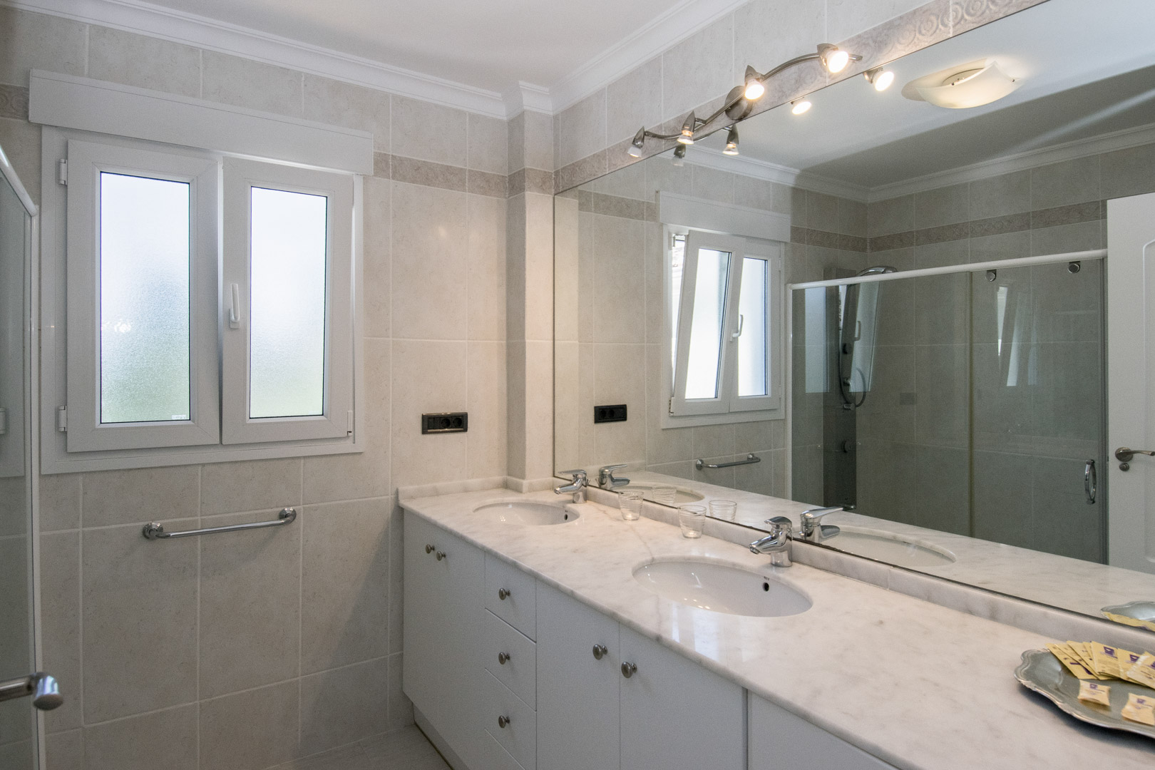 Badezimmer mit Doppelwaschtisch aus Marmor, weißen Unterschränken und großem Spiegel mit Beleuchtung. Neutrale Wandfliesen und ein mattiertes Dreh-Kipp-Fenster.