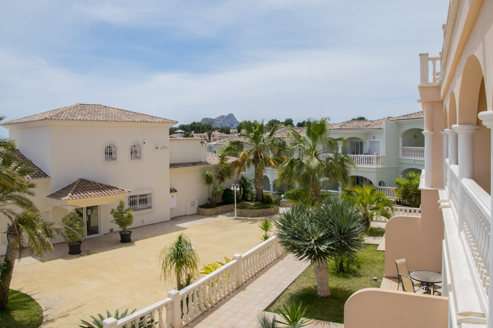 Blick von der Terrasse der Anlage auf die mediterrane Architektur, gepflegte Gärten mit Palmen und die Silhouette des Peñón de Ifach in der Ferne.