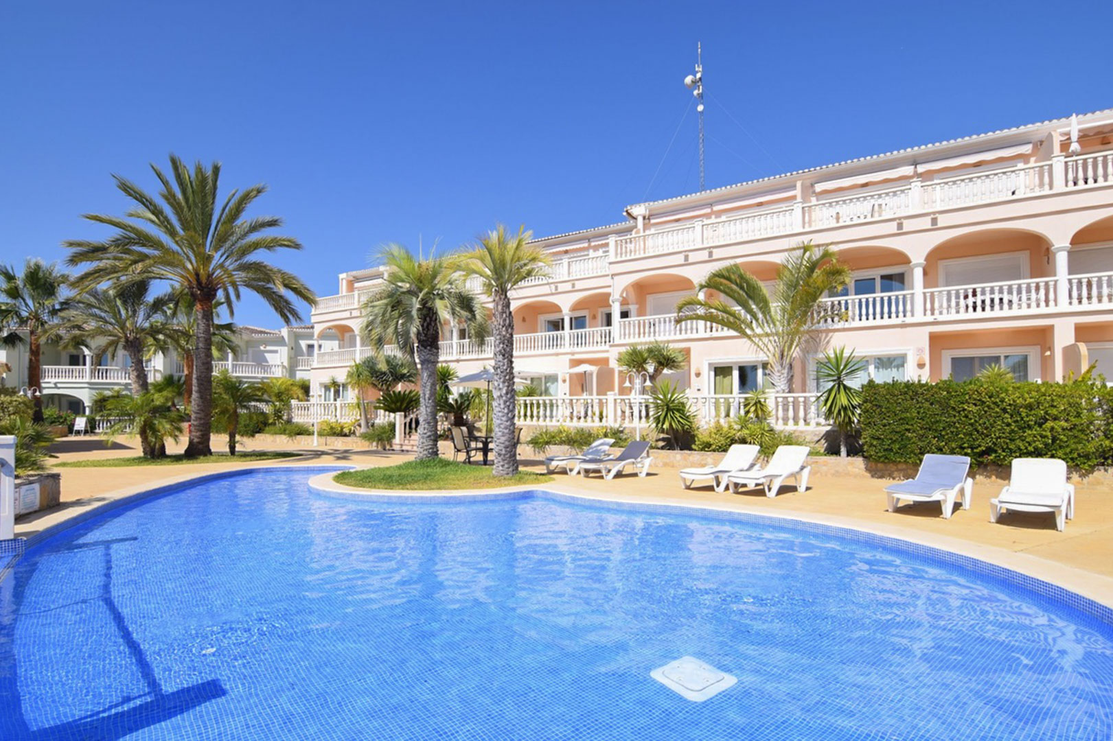 Gemeinschaftlicher Lagunenpool mit blauen Fliesen, umgeben von Palmen und Sonnenliegen vor einem mediterranen Gebäude mit bogenförmigen Terrassen und weißen Balustraden.