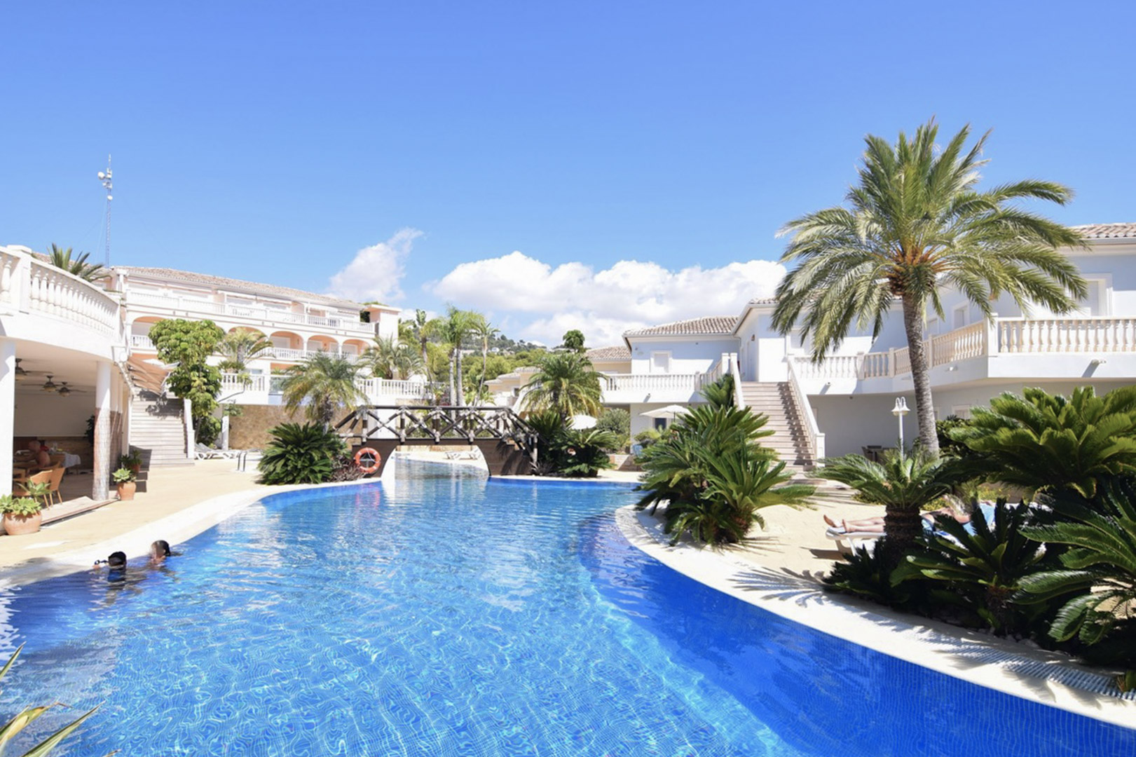 Gemeinschaftlicher Lagunenpool mit Holzbrücke, umgeben von Palmen und mediterraner Architektur mit weißen Balkonbalustraden in Benissa Costa.