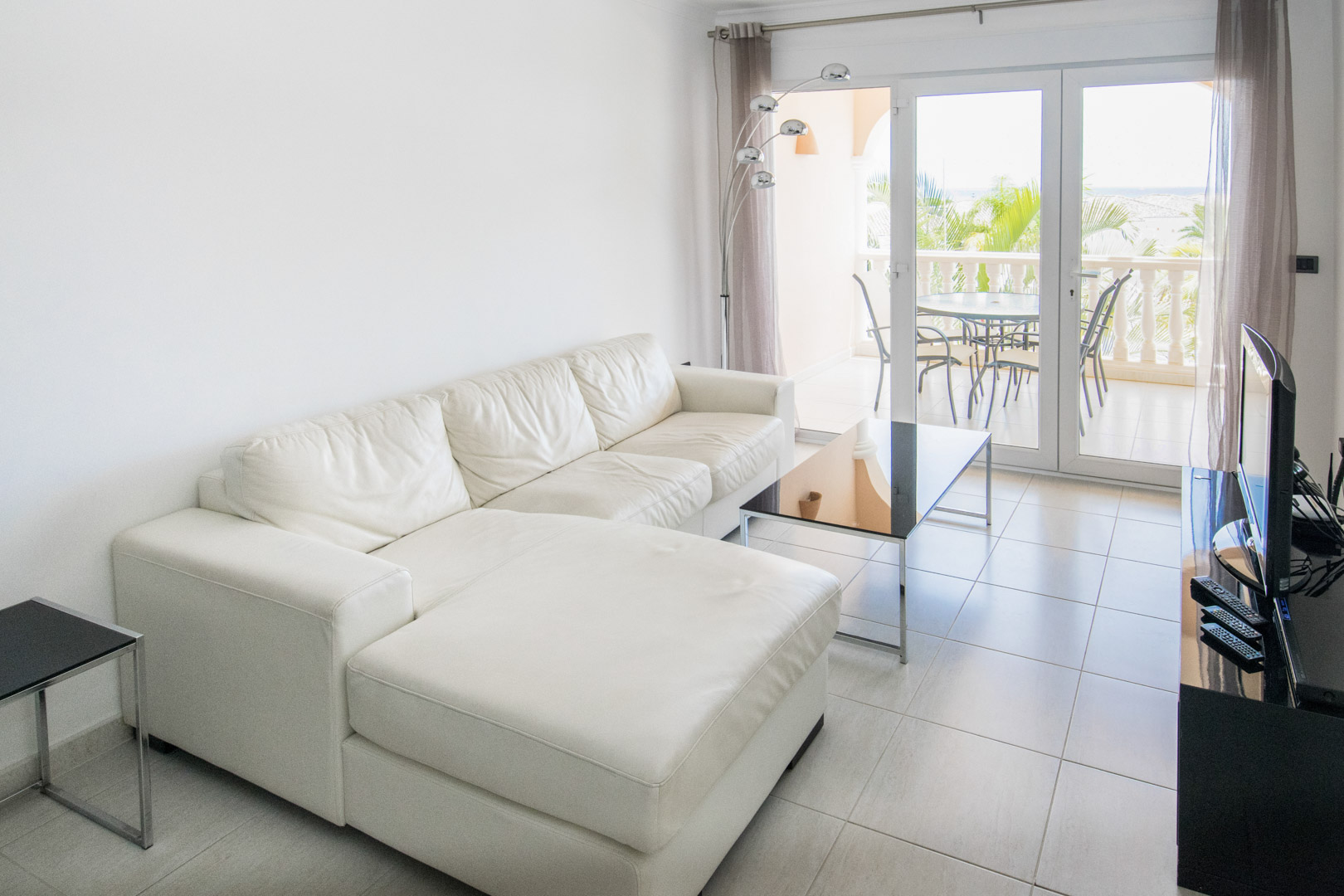 Heller Wohnbereich mit weißem Ledersofa, Fliesenboden und großen Glastüren, die auf eine möblierte Terrasse mit Blick auf Palmen und das Meer führen.