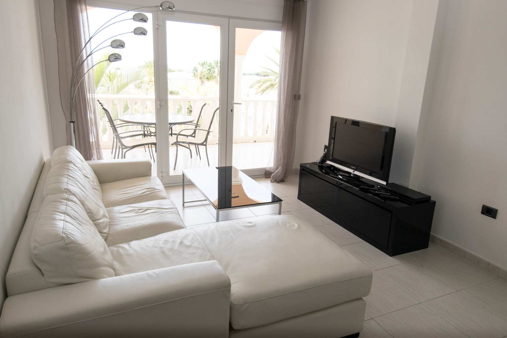 Heller Wohnbereich mit weißem Ledersofa, schwarzem Hochglanz-TV-Möbel und bodentiefen Glastüren, die auf eine Terrasse mit Palmenblick führen.