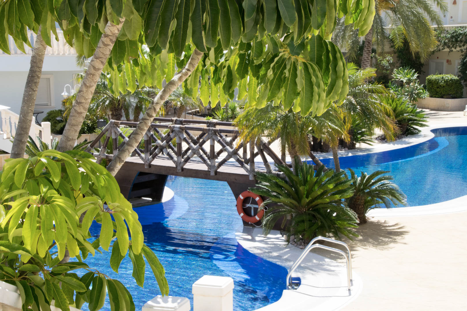 Lagunenartiger Pool mit dekorativer Holzbrücke, umgeben von üppigen mediterranen Gärten und tropischen Palmen im Außenbereich.