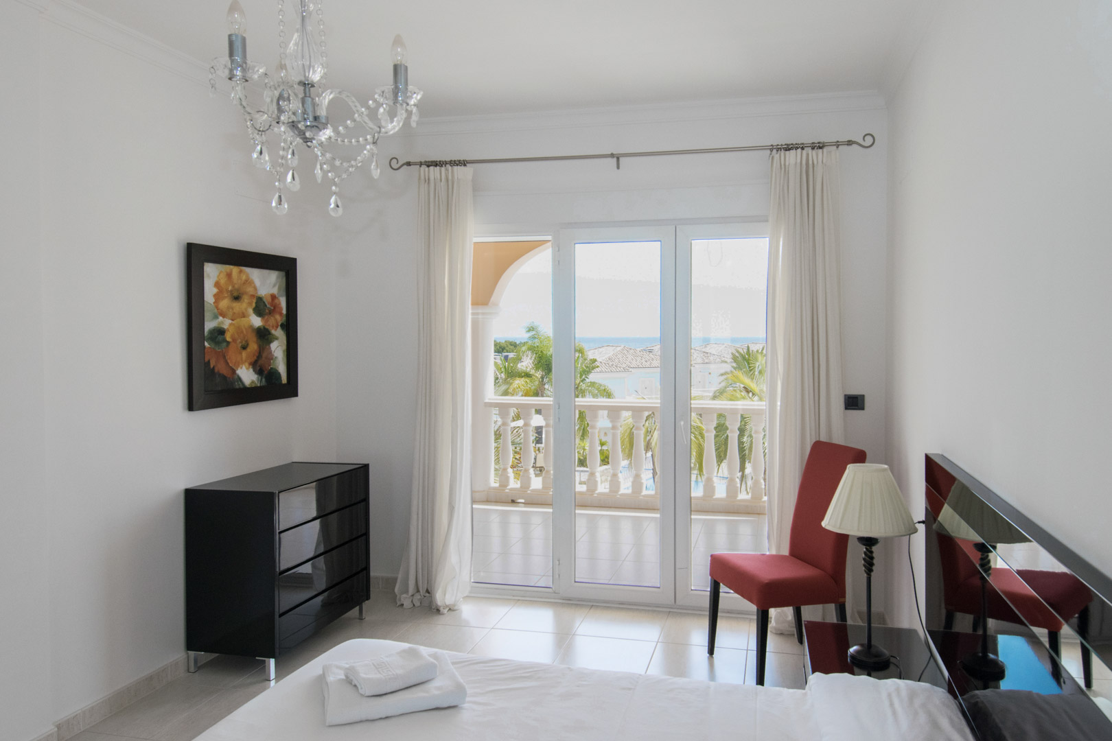 Schlafzimmer mit Terrassenzugang, moderner Kommode und Meerblick. Ausgestattet mit Kristallleuchter, hellen Fliesen und großen Glastüren zum privaten Balkon.