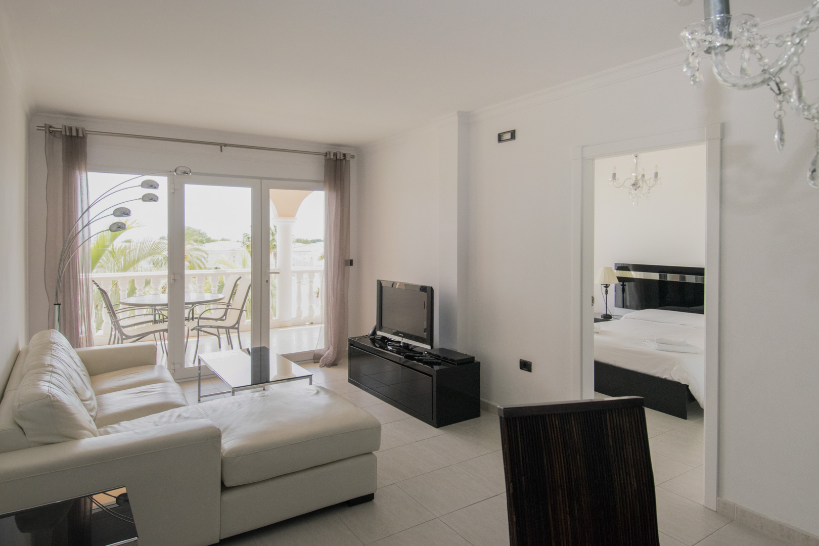 Wohnzimmer mit weißem Ledersofa, schwarzen Designmöbeln und Terrassenzugang. Der Raum besticht durch Kristallleuchter und einen offenen Übergang zum Schlafzimmer.