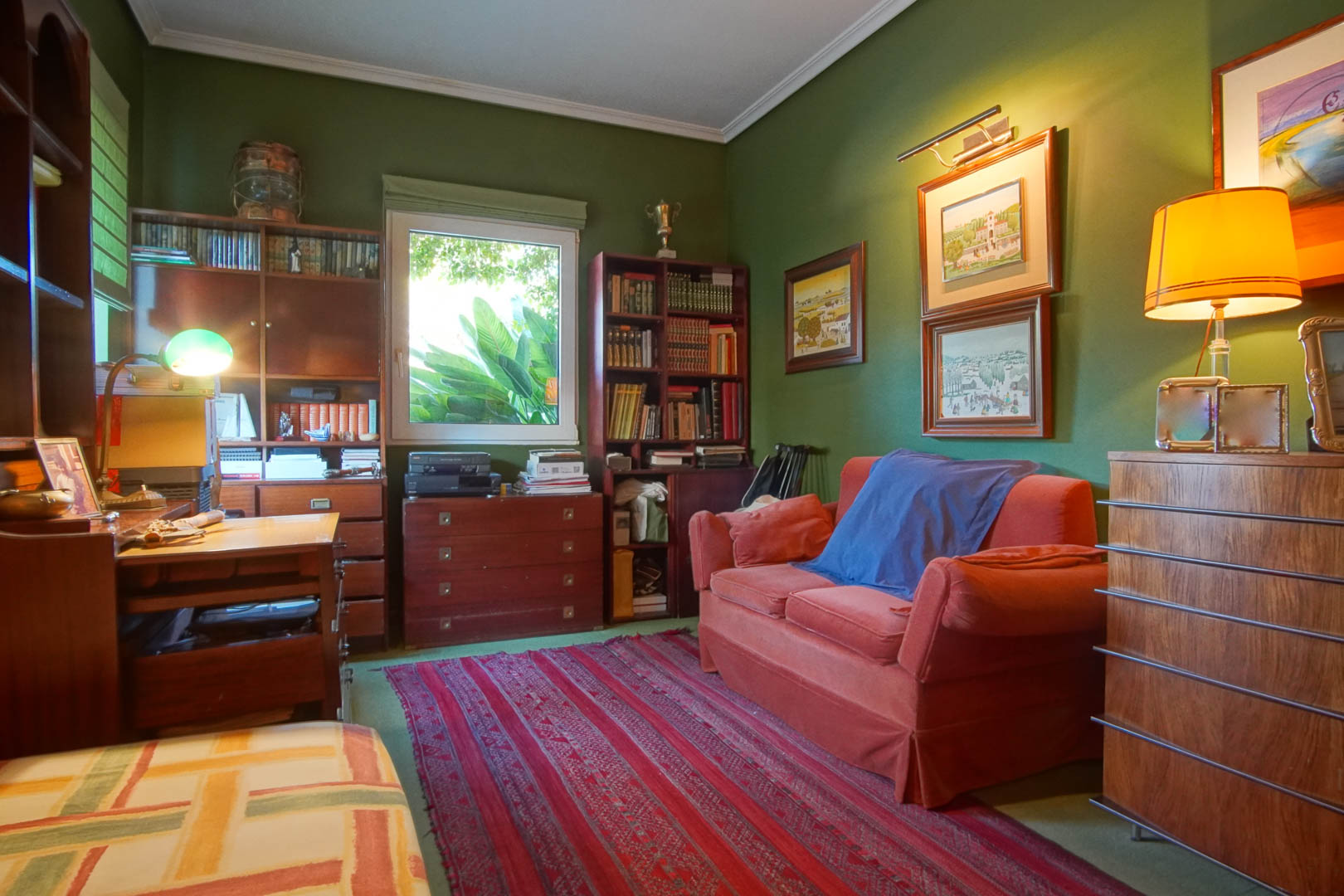 Arbeitszimmer mit grünen Wänden und klassischen Holzmöbeln. Es bietet Bücherregale, einen Schreibtisch, ein rotes Sofa und Tageslicht mit Blick ins Grüne.