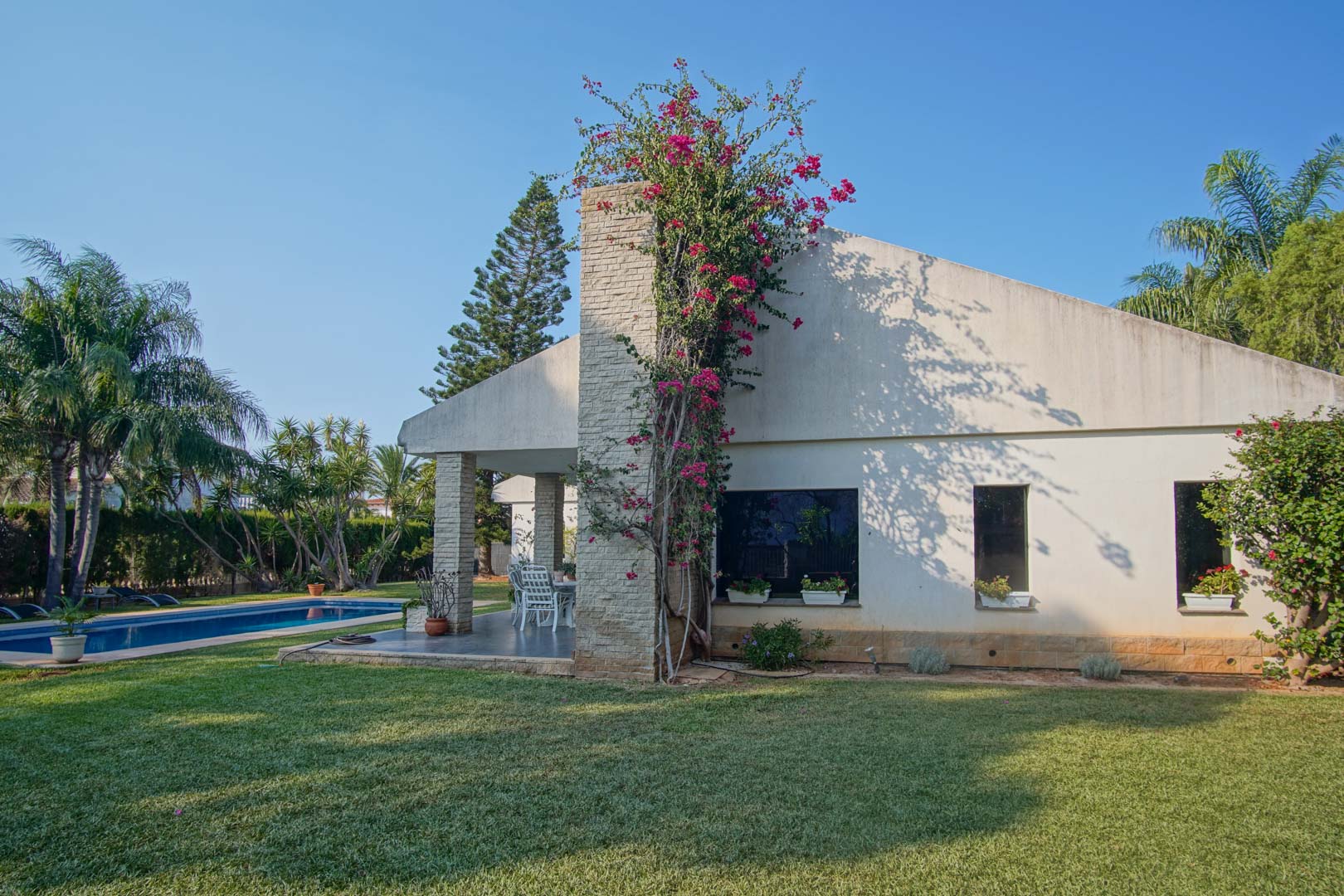 Außenfassade mit steinverkleidetem Kamin und Bougainvillea, gepflegter Rasenfläche und privatem Pool in einer mediterranen Gartenanlage.