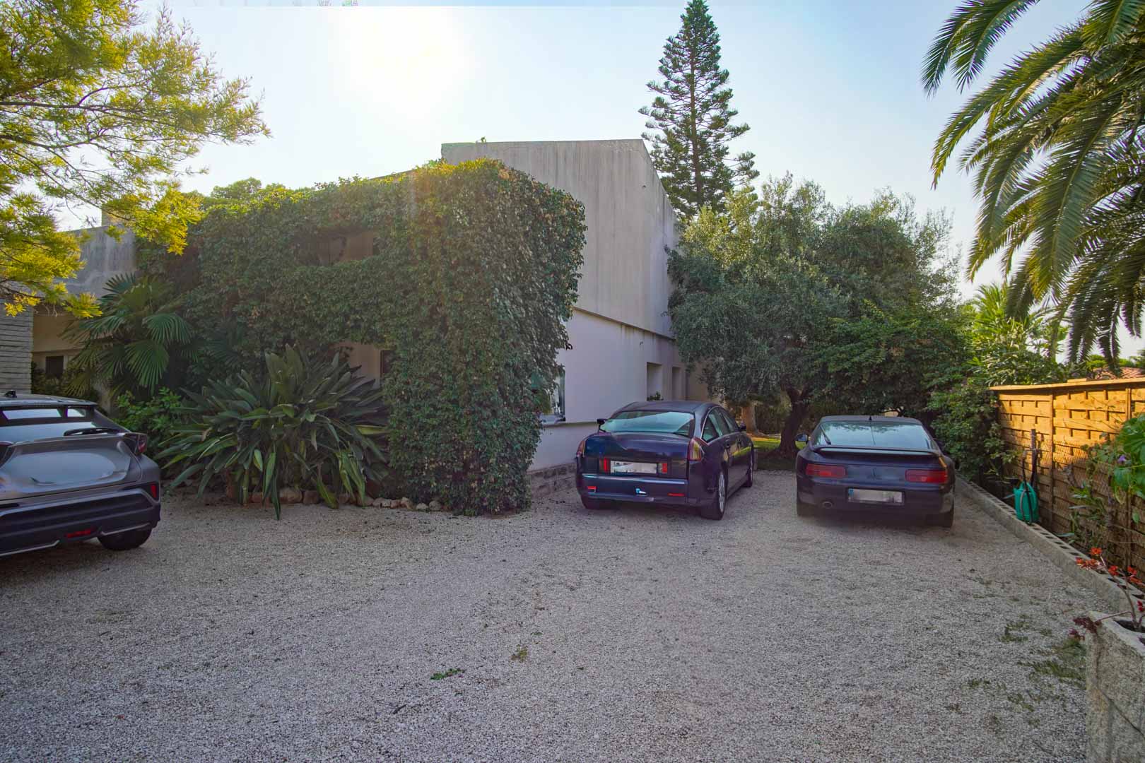 Außenparkplatz mit Kiesoberfläche, umgeben von mediterraner Vegetation und einem Holzzaun. Der Bereich bietet Platz für mehrere Fahrzeuge auf dem Privatgrundstück.