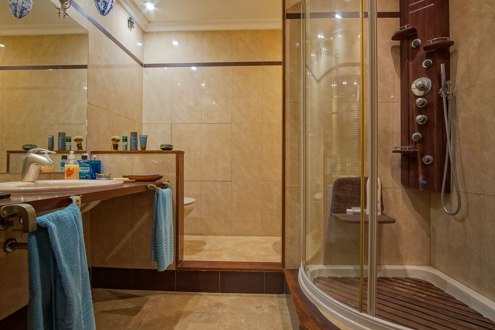 Badezimmer mit beigen Marmorfliesen, Holzwaschtisch und einer runden Duschkabine mit Hydromassage-Säule und Holzlattenrost.