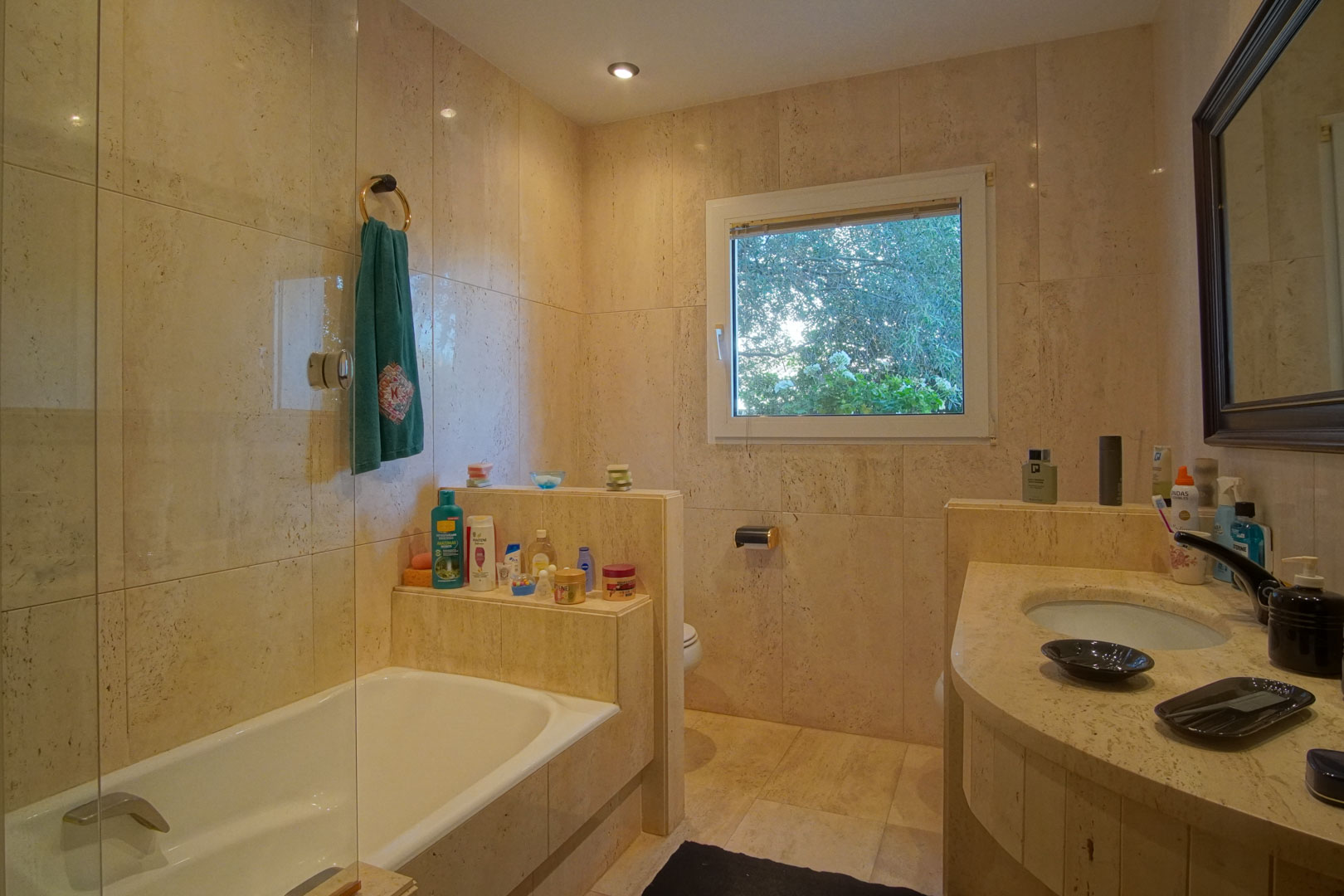 Badezimmer mit cremefarbener Marmorverkleidung, Einbaubadewanne mit Glaswand und Naturstein-Waschtisch. Ein Außenfenster sorgt für Tageslicht und Gartenblick.
