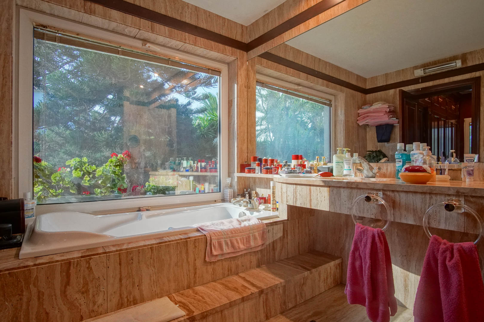 Badezimmer mit Travertin-Marmor-Verkleidung, Einbaubadewanne an einem großen Fenster mit Gartenblick und integriertem Waschtisch mit großem Spiegel.