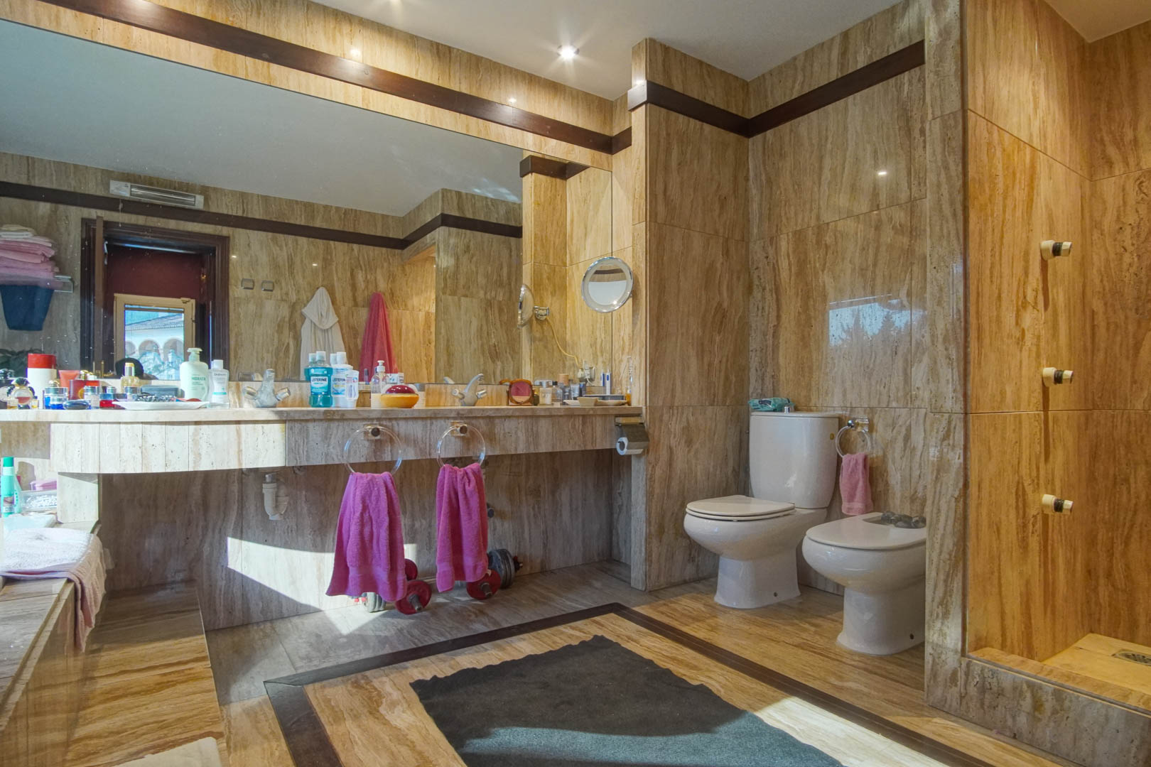 Badezimmer mit vollständiger Travertin-Marmorverkleidung, integriertem Waschtisch, großem Spiegel und Einbaustrahlern für ein elegantes Ambiente.