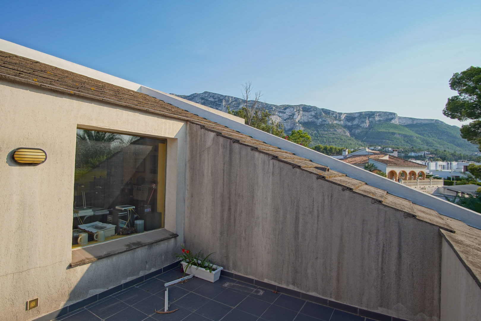 Balkon im Obergeschoss mit dunklen Bodenfliesen und freiem Blick auf das Montgó-Massiv. Das große Fenster bietet Einblick in den angrenzenden Bürobereich.