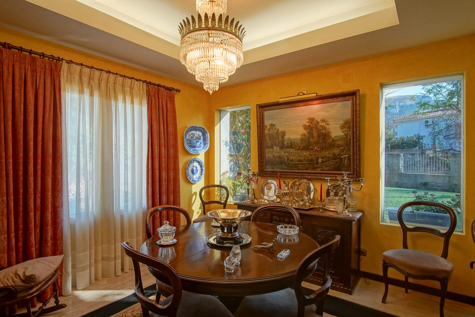 Elegantes Esszimmer mit rundem Holztisch, Kristallleuchter und klassischem Sideboard. Ockerfarbene Wände, eine Lichtvoute und der Blick in den Garten prägen das Ambiente.