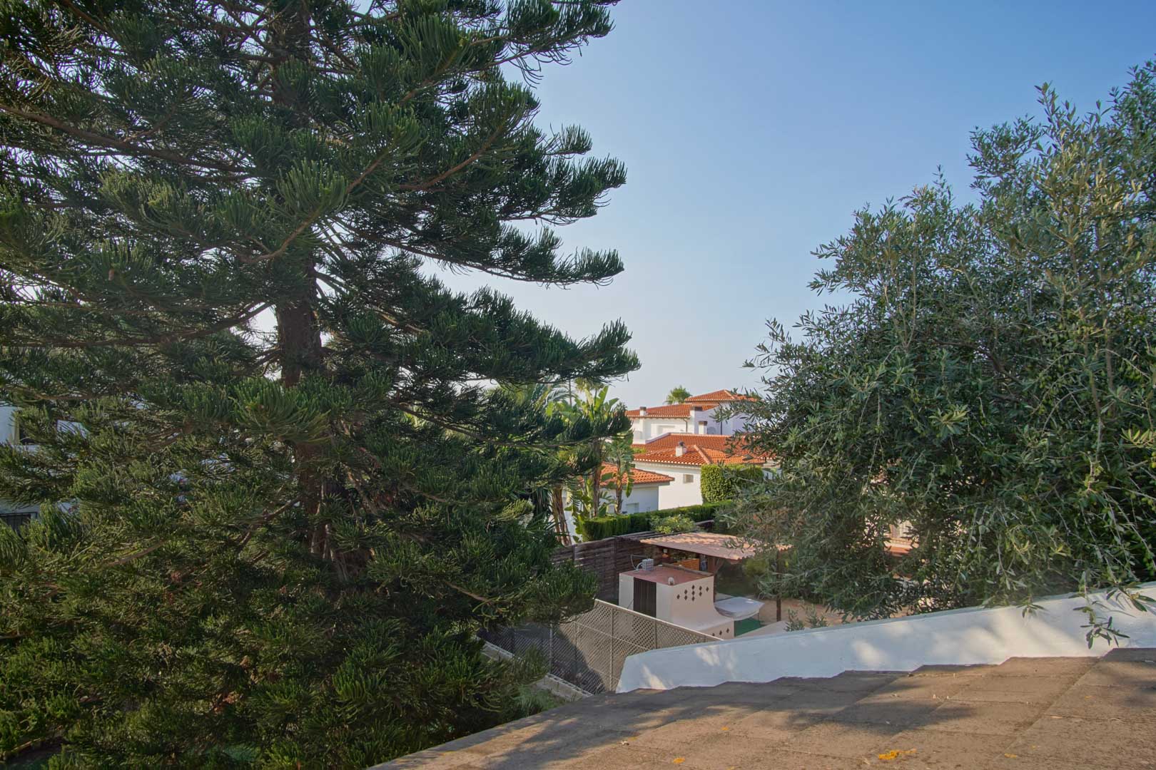 Erhöhter Blick von der Terrasse auf die mediterrane Vegetation und die Terrakotta-Dächer der benachbarten Villen unter klarem Himmel.