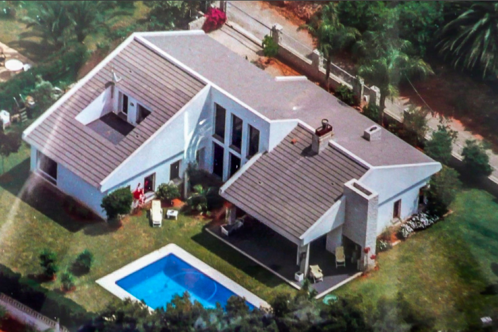 Luftaufnahme der weißen Villa mit Ziegeldach, rechteckigem Pool und gepflegtem Garten mit überdachter Terrasse und Umzäunung.