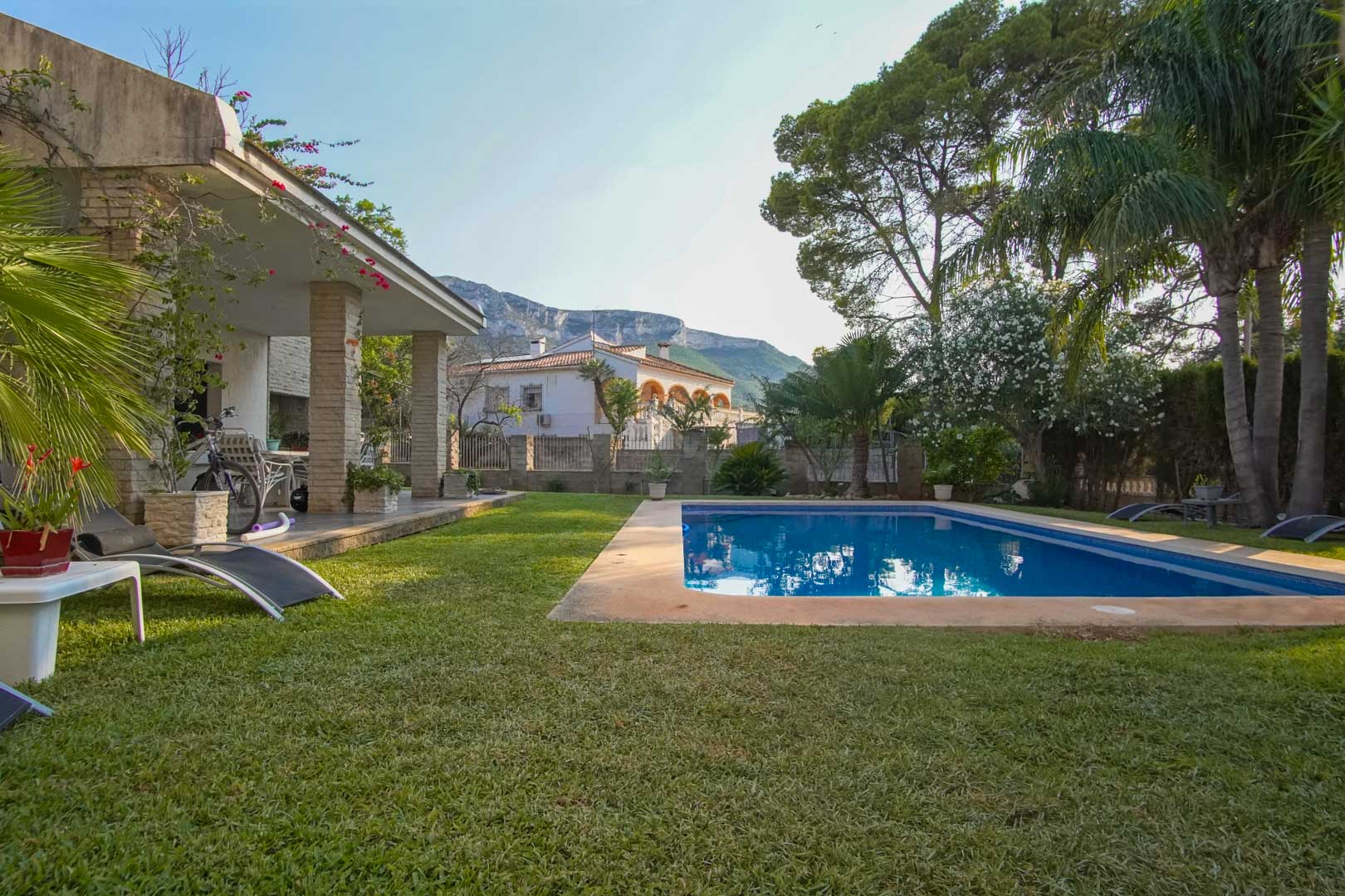 Mediterraner Garten mit Naturrasen, rechteckigem Pool und überdachter Terrasse mit Steinsäulen. Im Hintergrund bietet der Berg Montgó eine markante Kulisse.