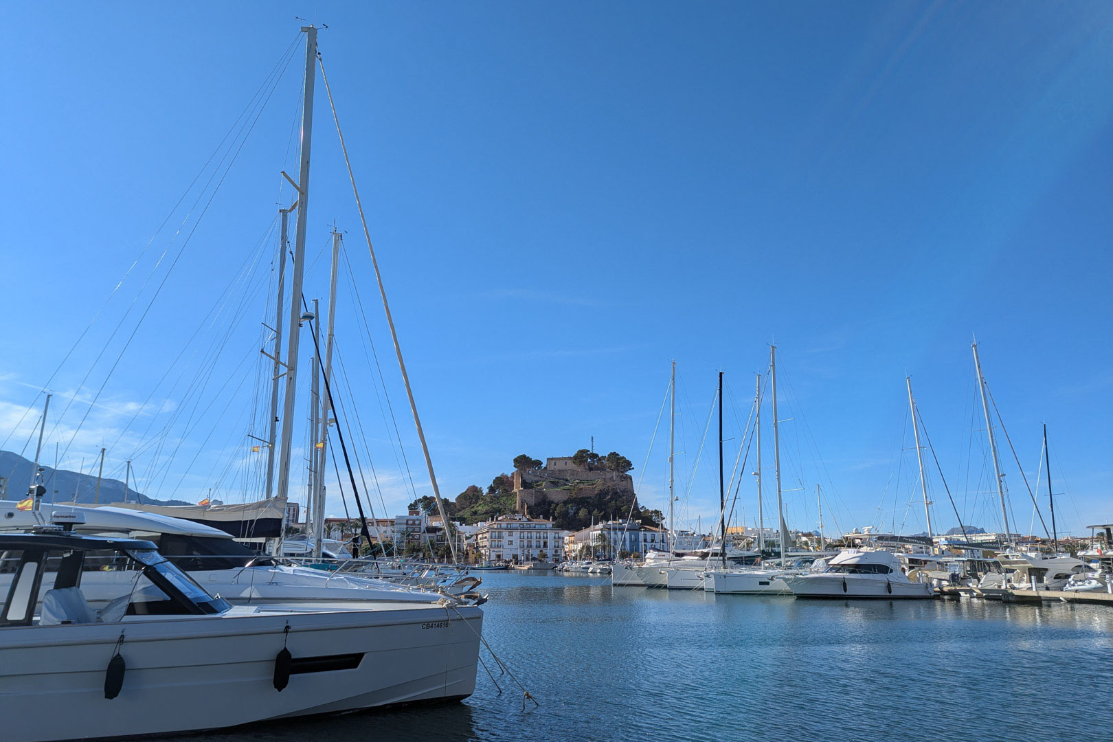 Panoramablick auf den Jachthafen von Dénia mit Segelbooten und der historischen Burg, die über der Stadt unter klarem blauem Himmel thront.