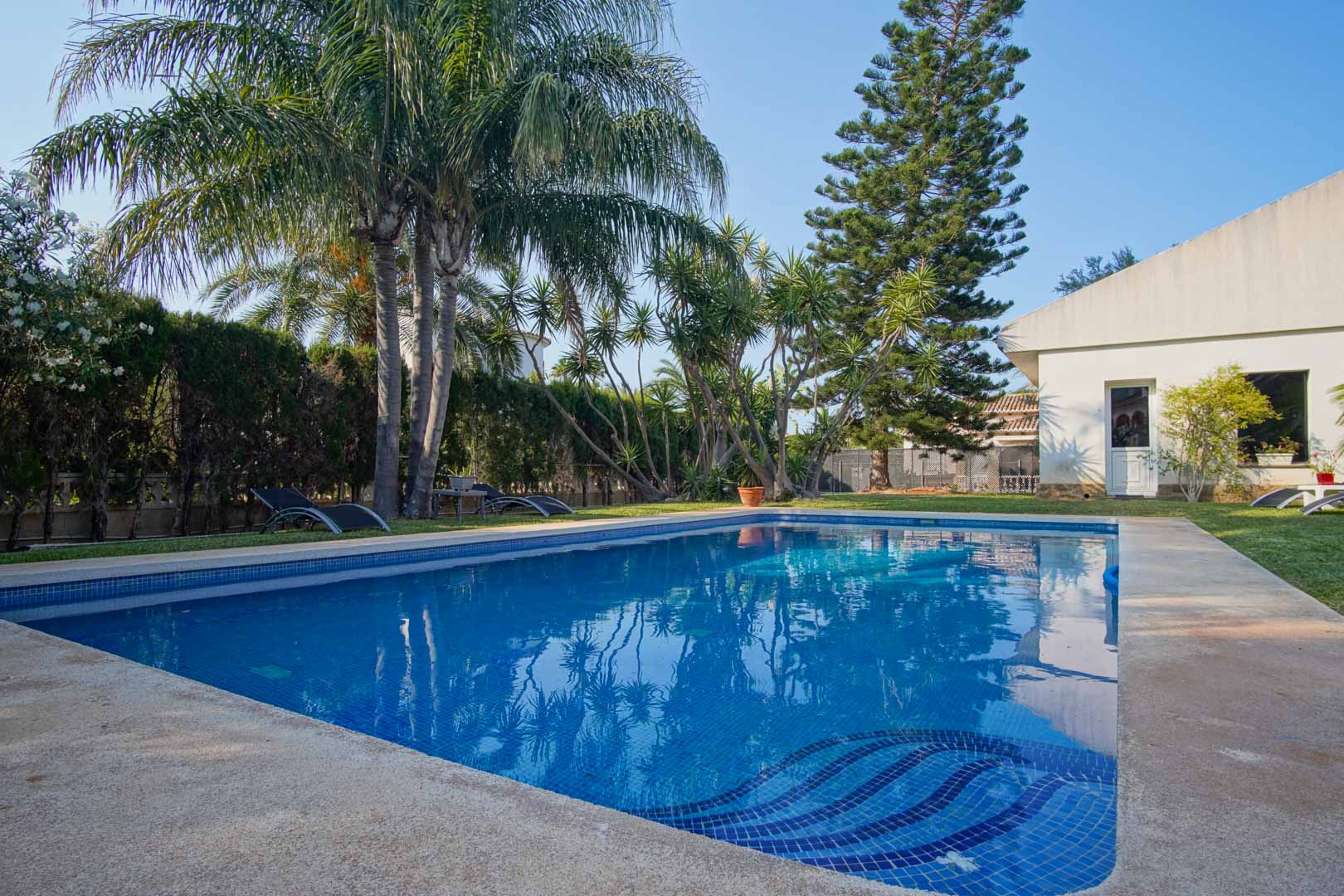 Privater Pool mit blauen Mosaikfliesen und integrierter Treppe, eingebettet in einen mediterranen Garten mit Palmen, Naturrasen und blickdichten Hecken.