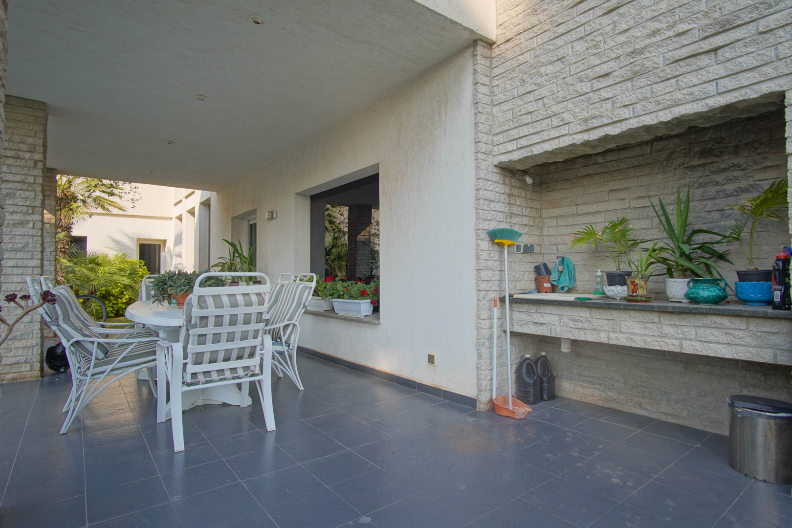 Überdachte Terrasse mit dunklen Bodenfliesen, Essbereich im Freien und integrierter Außenküche aus Naturstein mit Spülbecken und Arbeitsplatte.