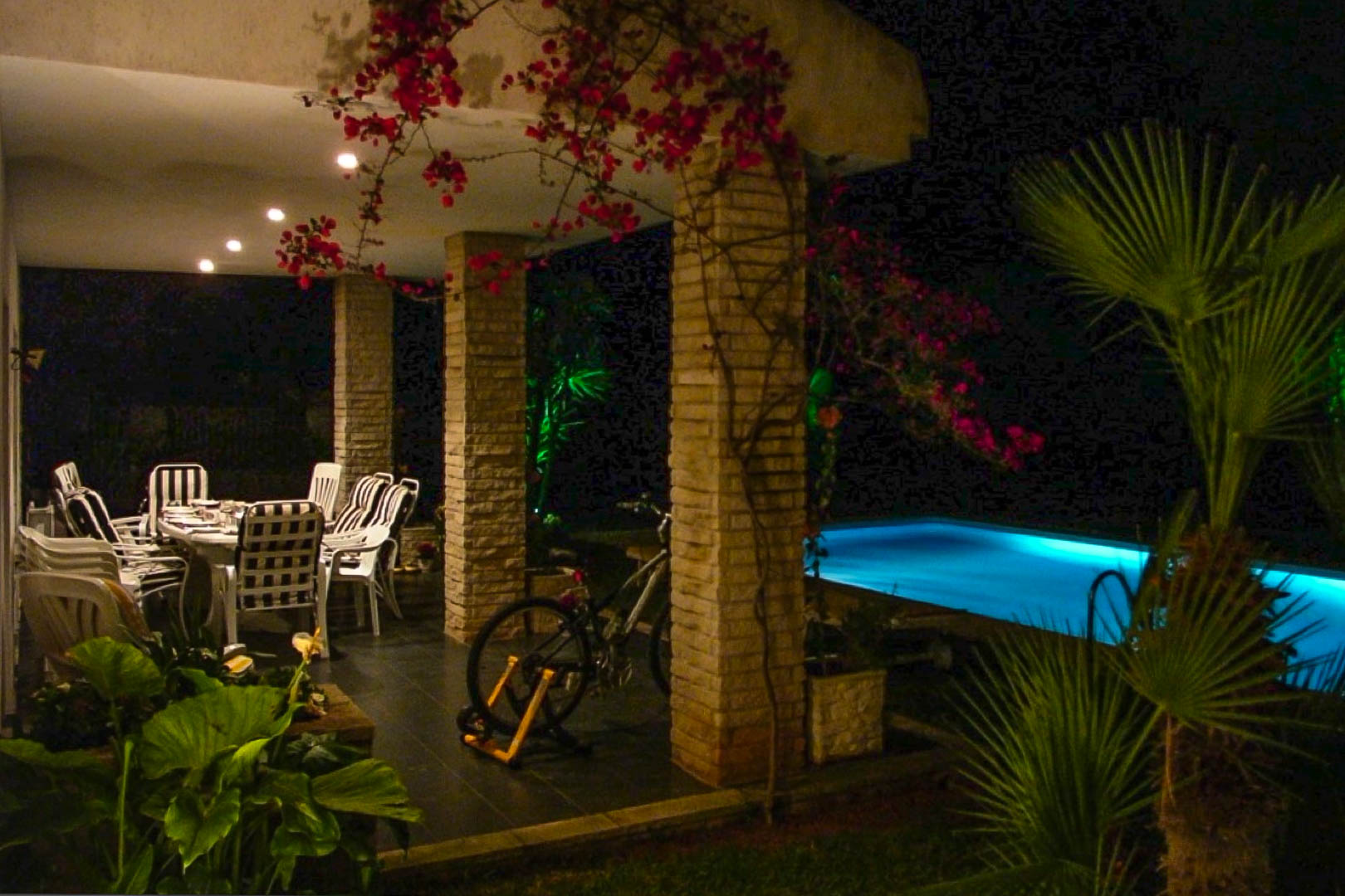 Überdachte Terrasse mit gemauerten Backsteinsäulen, Essbereich im Freien und Bougainvillea neben einem nachts beleuchteten Swimmingpool.